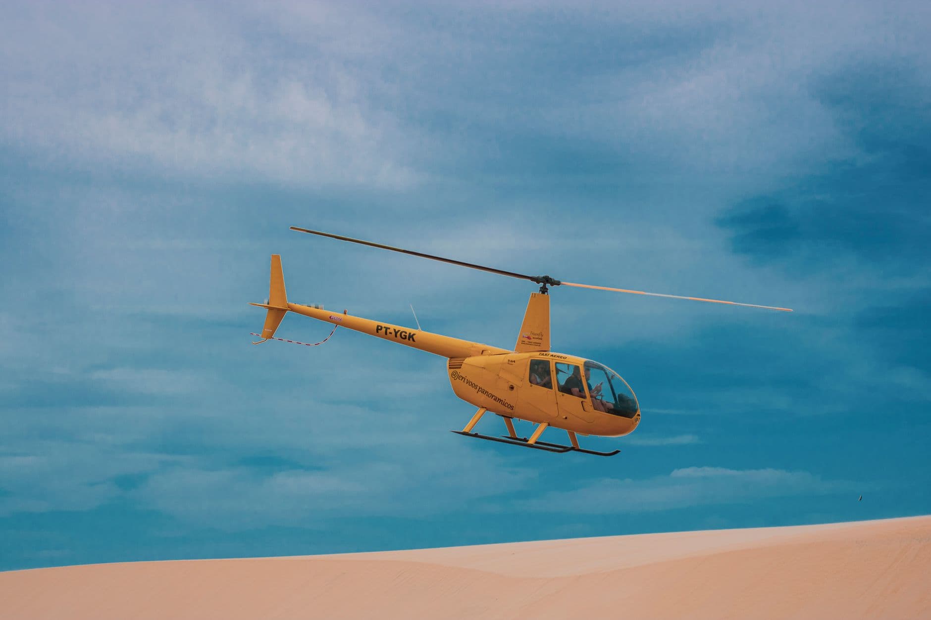 A vibrant yellow helicopter soars over the vast dunes of Vila de Jericoacoara under a clear blue sky.