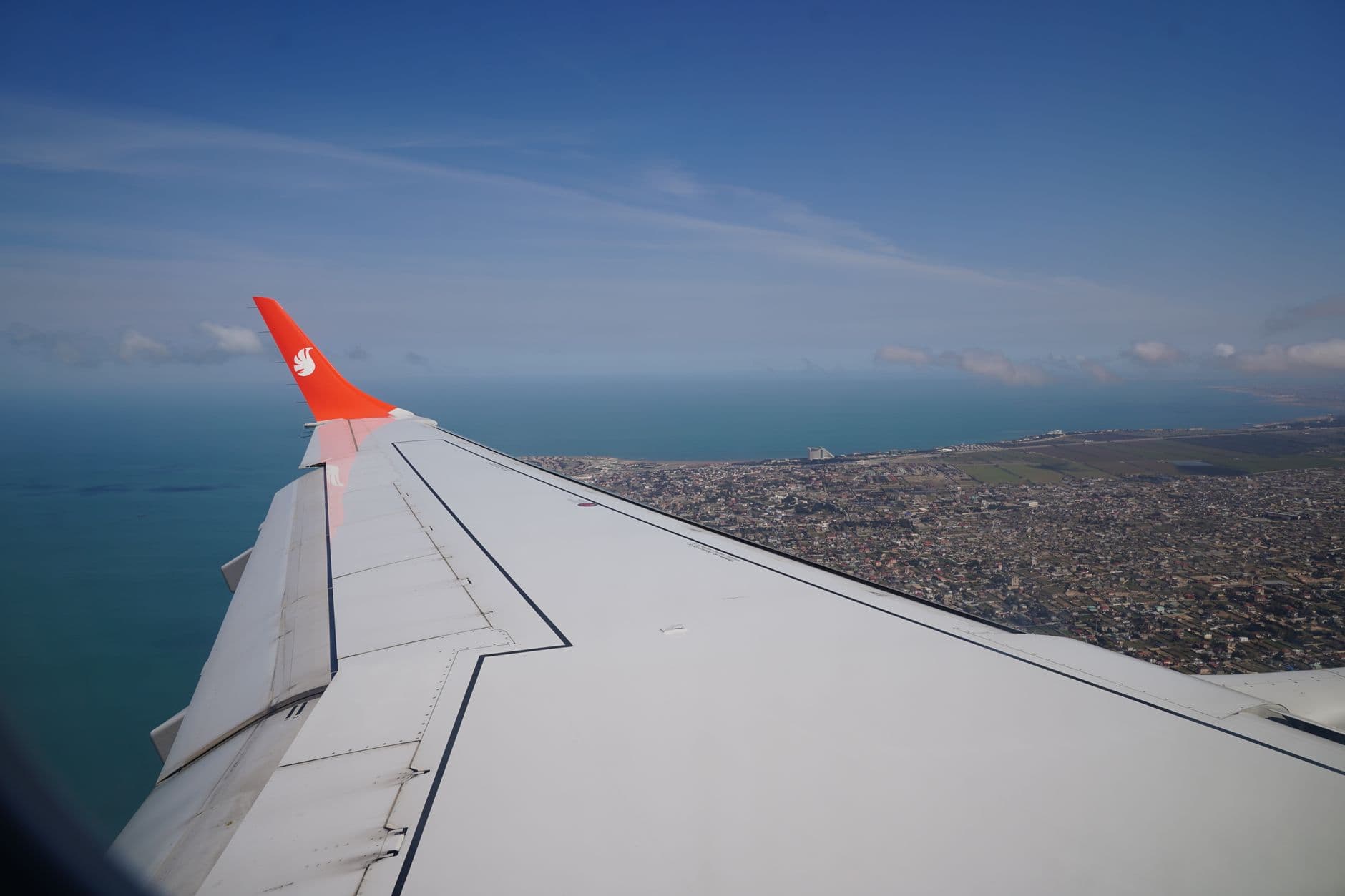 View from airplane window showing wing and coastal city below with vast ocean under blue sky.
