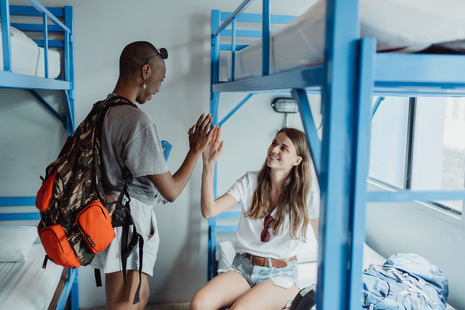 Two friends with a backpack high-fiving in a bright hostel room with blue bunk beds.