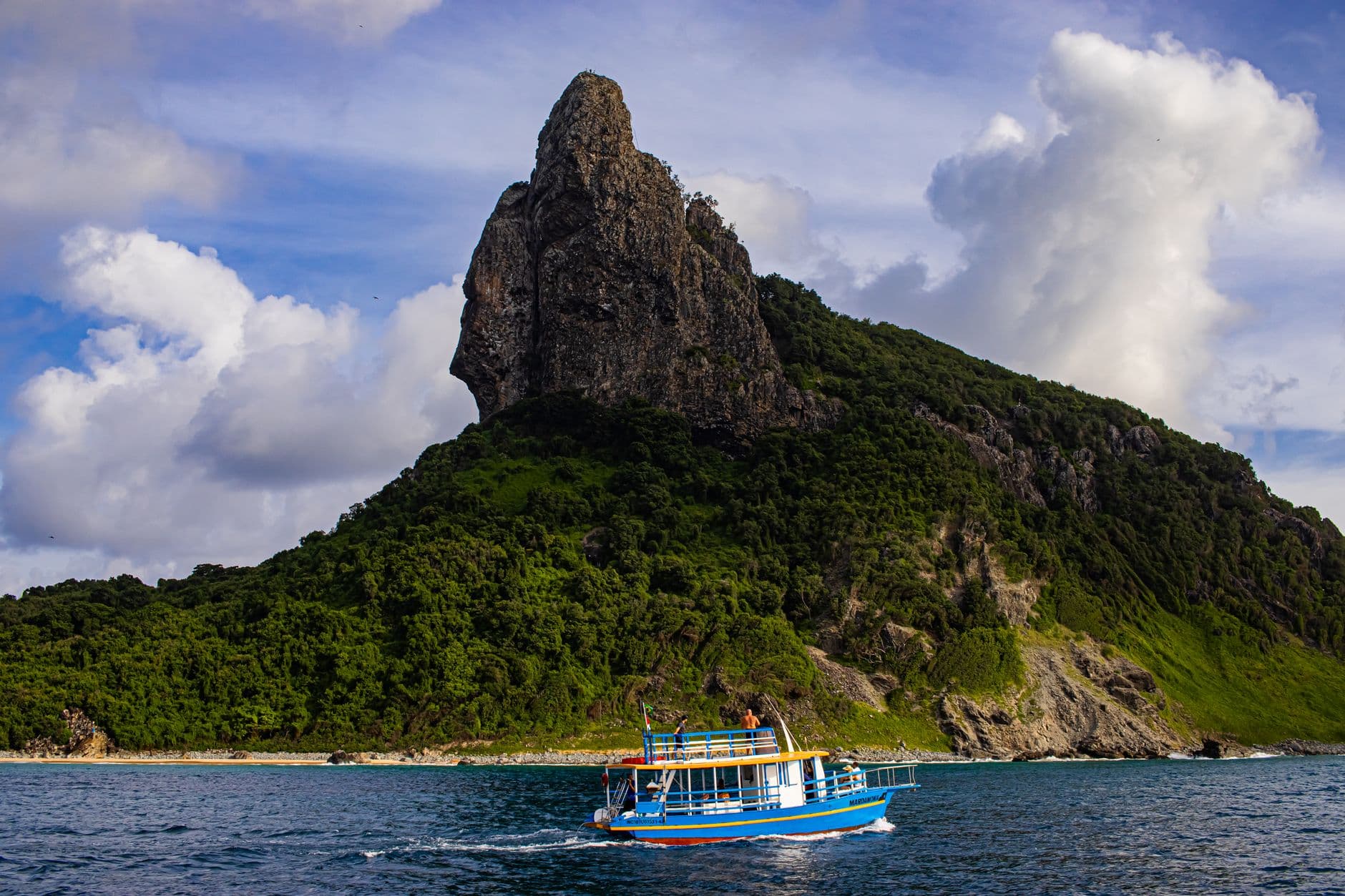 A vibrant boat sails by a stunning rocky island with lush greenery under a dramatic sky.