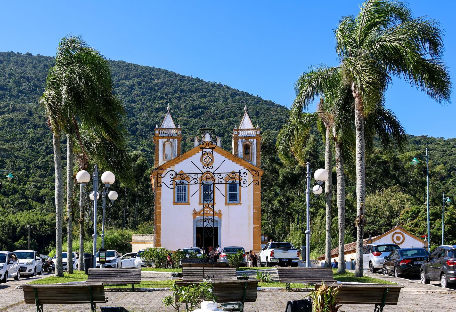 Colonial-style church with palm trees and cars, set against a forested hill under a blue sky.
