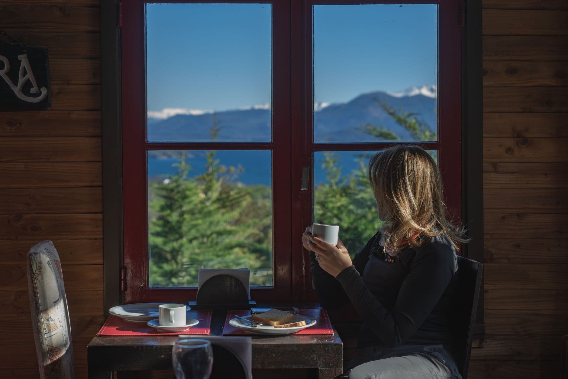 A woman enjoys coffee indoors with a scenic mountain view in Bariloche, Argentina.