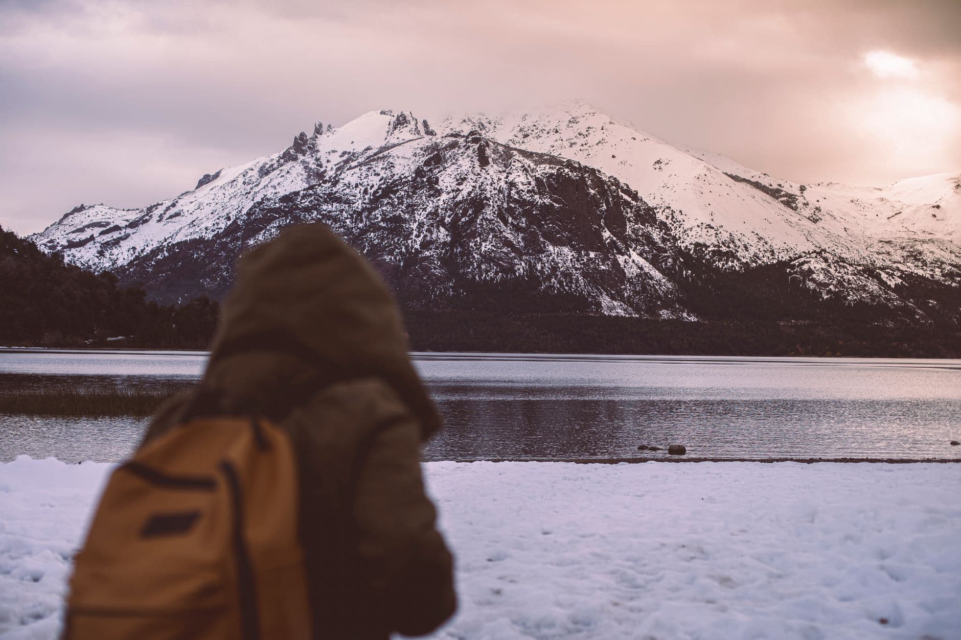 Adventurer views snow-capped mountains by a serene Patagonian lake during winter sunset.