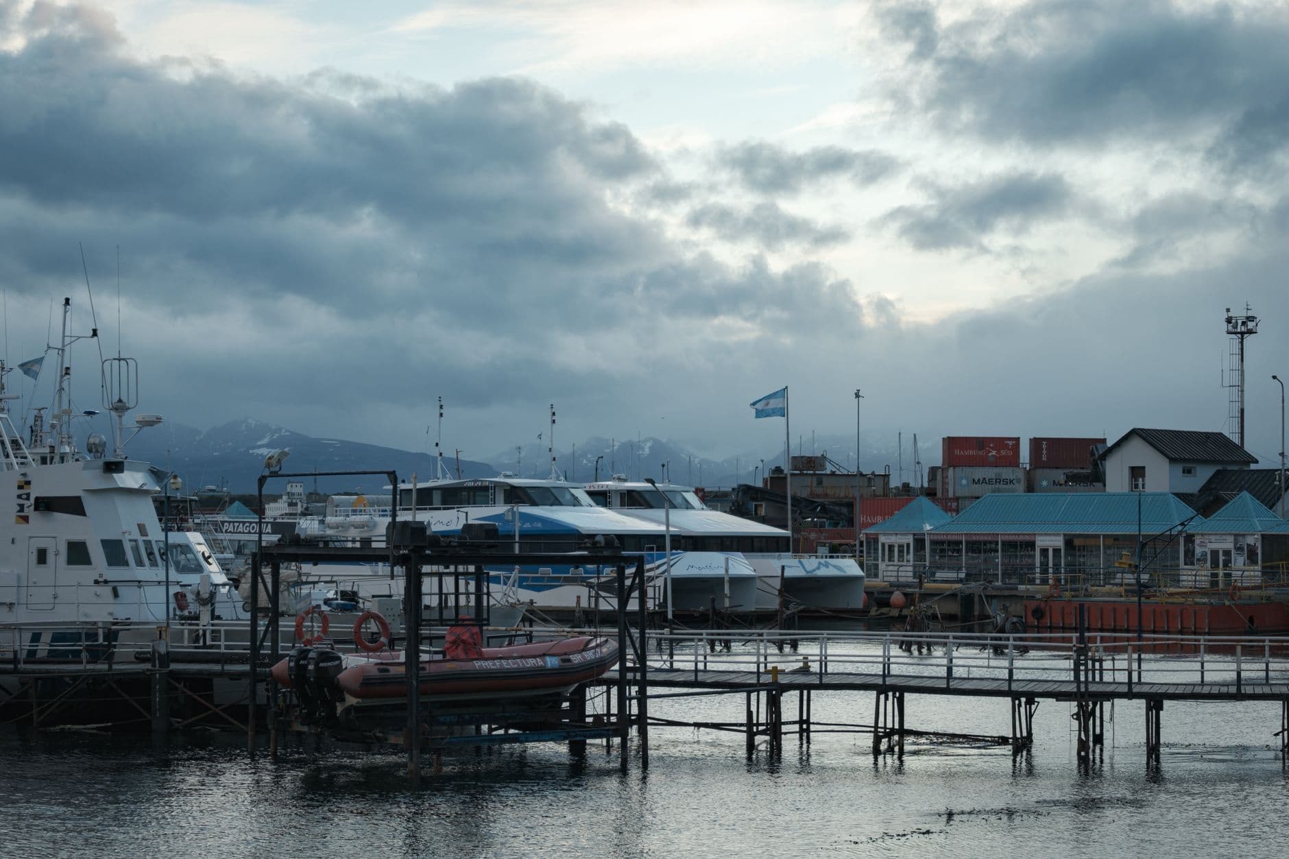 Scenic view of docked boats at Ushuaia harbor with mountain backdrop and cloudy sky.