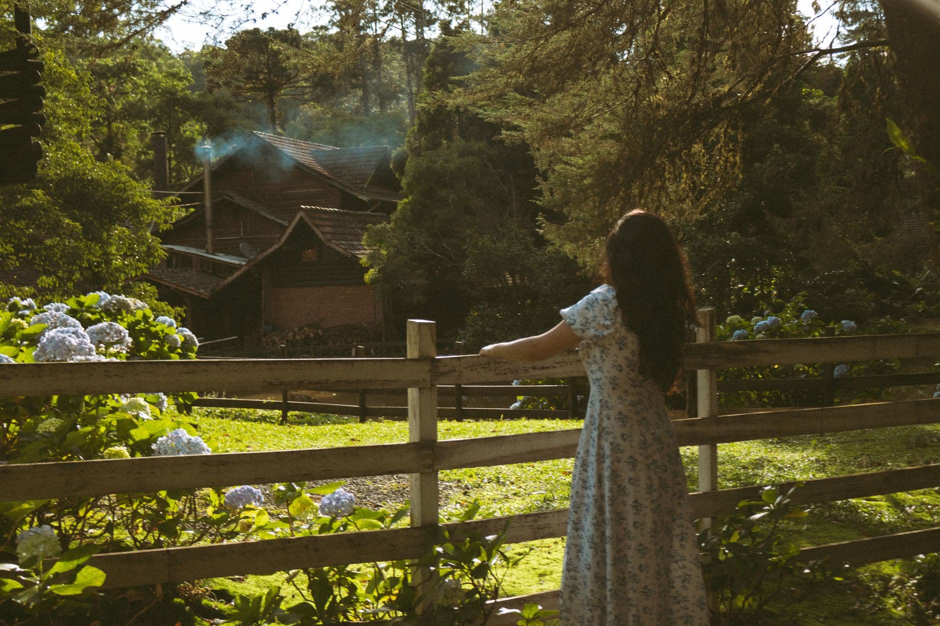 Woman in a floral dress views a serene countryside house from a wooden fence.