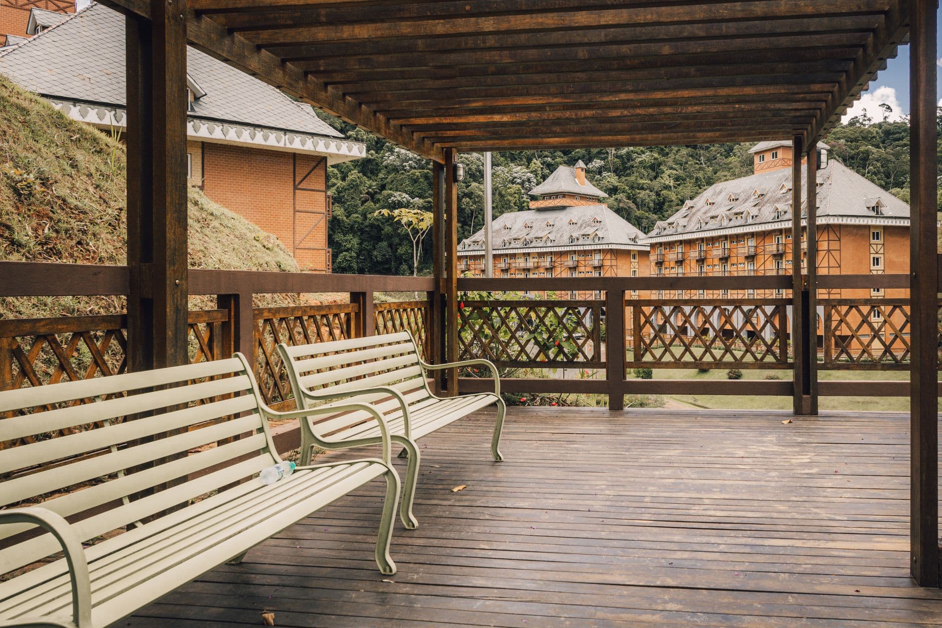A scenic wooden terrace with benches faces a charming rustic building amidst lush greenery.