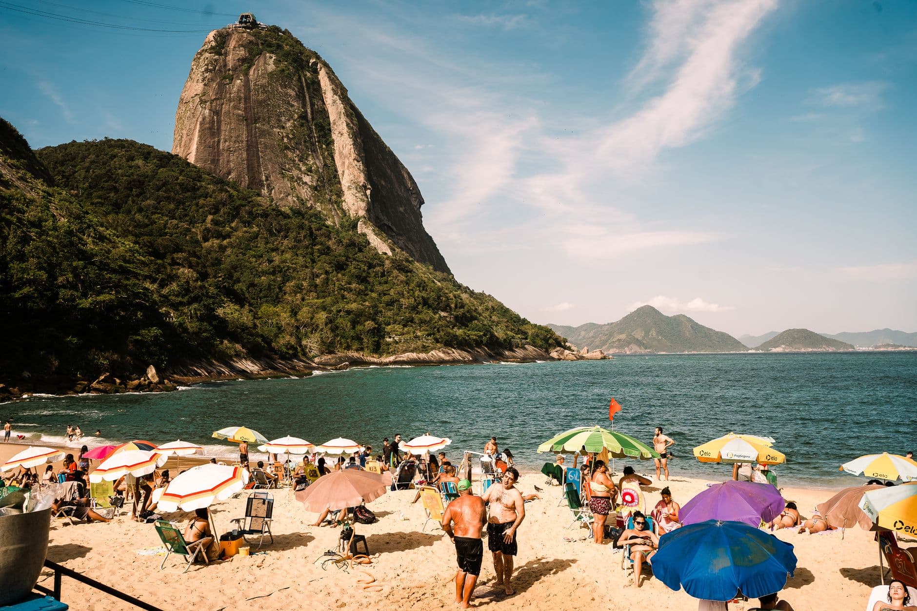Crowded beach scene under Sugarloaf Mountain in Rio, with vibrant umbrellas and lively atmosphere.